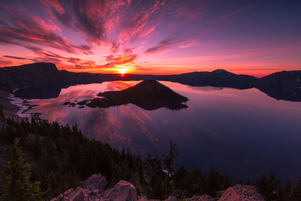 A sunrise over Crater Lake paints the sky and reflections in the water with brilliant pinks and purples with scattered clouds texturing the sky.