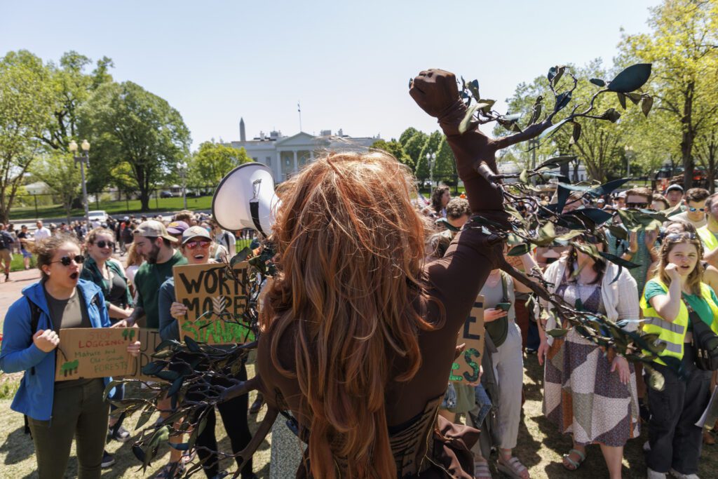 A crowd with hand-made signs gathers in front of the White House. Pattie Gonia leads a chant into a megaphone.