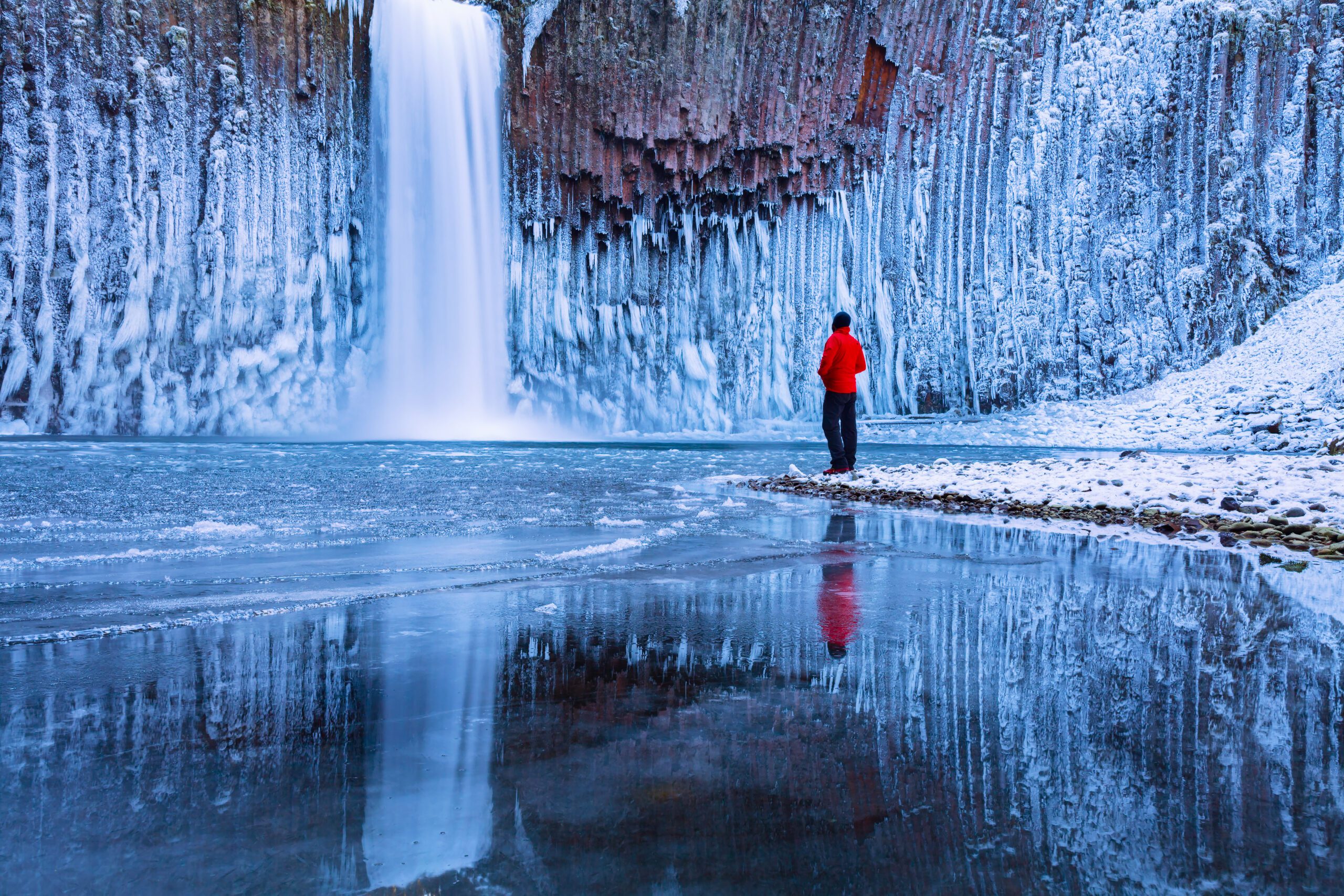 A man in a red jacket stands looking at a frozen waterfall and the ice-covered rocks behind it