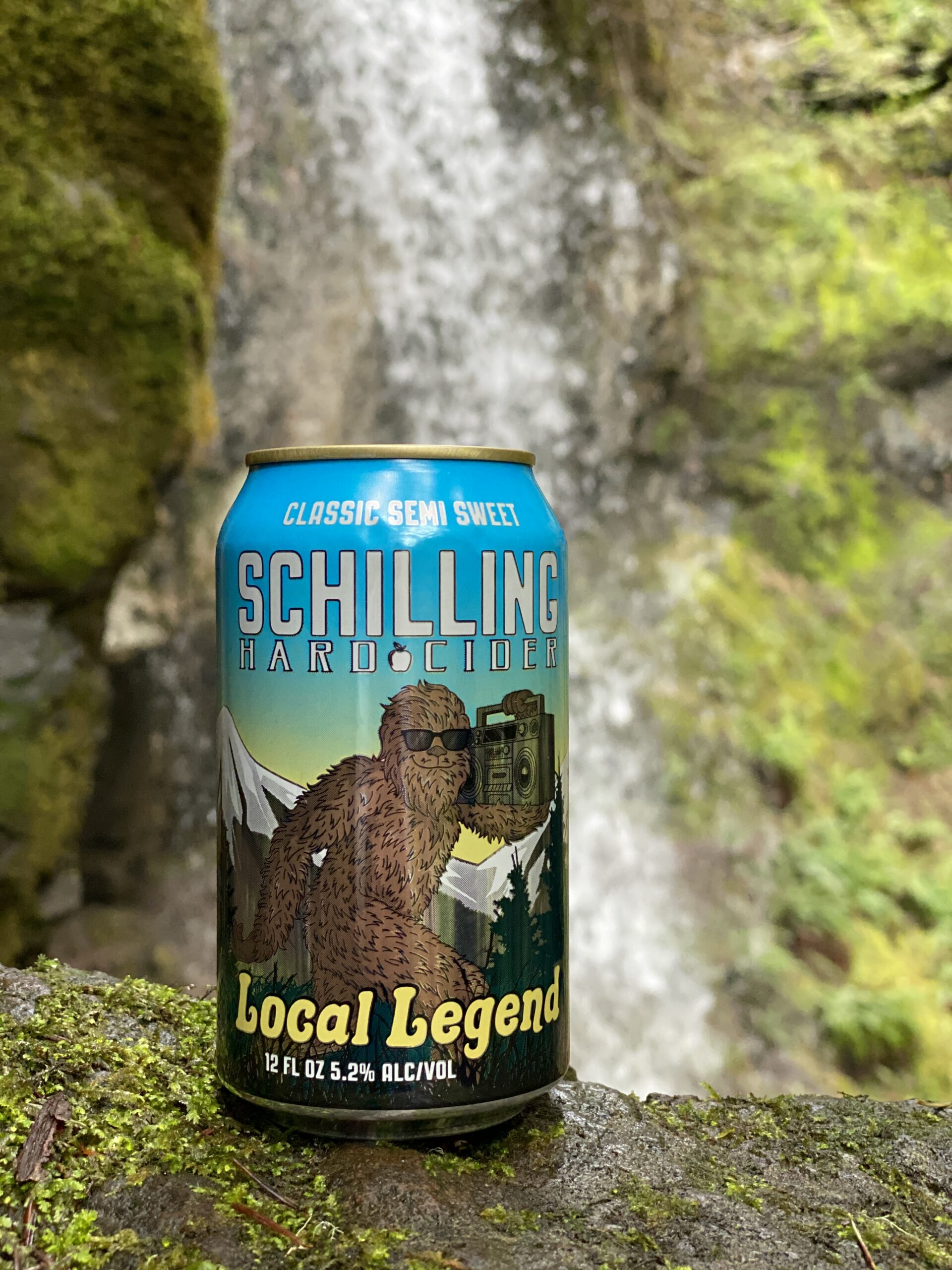 A can of Schilling Cider's Local Legend featuring a sasquatch in sunglasses sits on a mossy rock in front of an out-of-focus waterfall.