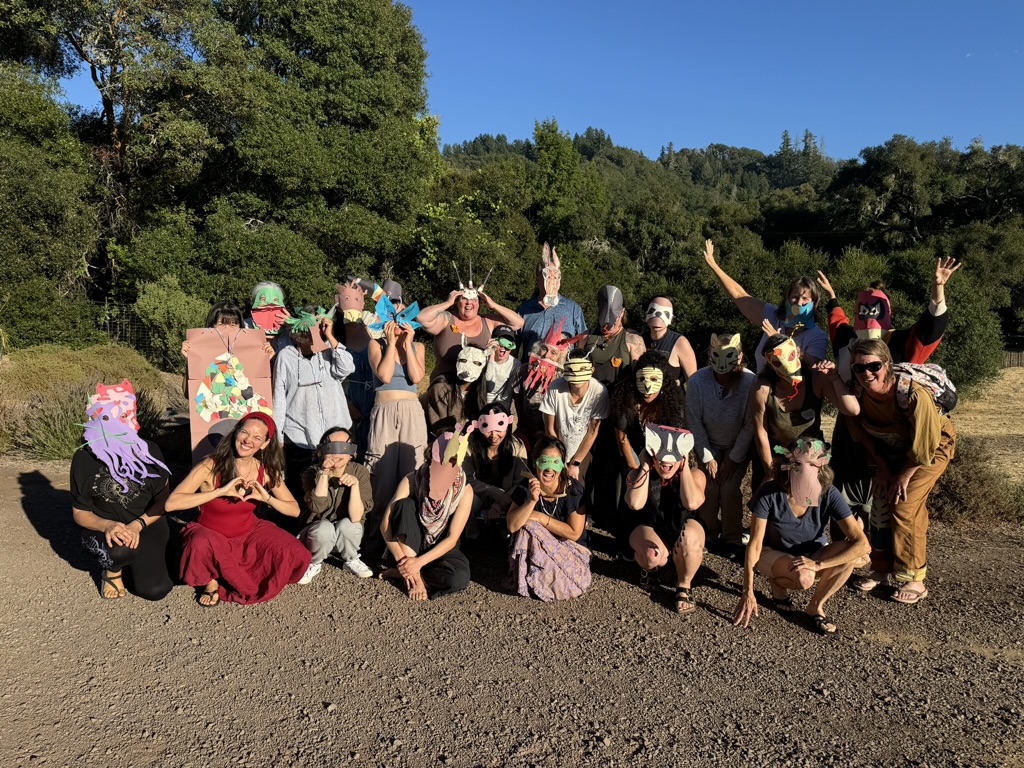 Image features a small group of smiling people outside in front of trees, most wearing costume masks